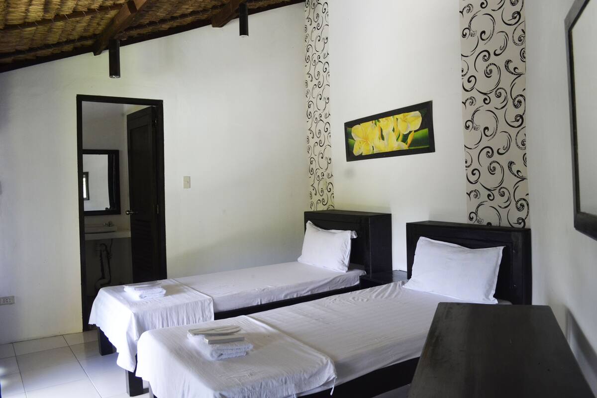 A room with two single beds, a wooden table, a mirror, a patterned curtain, and a framed picture on the wall.