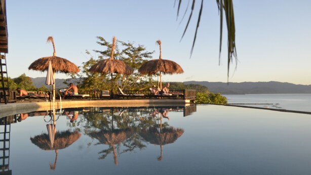Una piscina infinita con sombrillas de paja y sillas de descanso, que refleja el paisaje circundante y las palmeras.