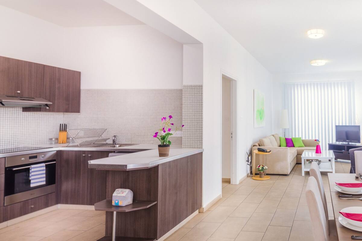 Una cucina moderna con forno integrato, un'area pranzo con tavolo apparecchiato per due e un soggiorno con divano e televisione.