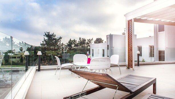 Una terraza moderna al aire libre con barandilla de vidrio, sillas blancas, una tumbona de madera y una mesa de vidrio.