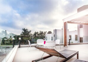 Una terraza moderna al aire libre con barandilla de vidrio, sillas blancas, una tumbona de madera y una mesa de vidrio.