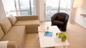 Una sala de estar moderna con un sofá color beige, un sillón oscuro, una mesa de centro blanca con un libro, una taza y una planta en maceta.
