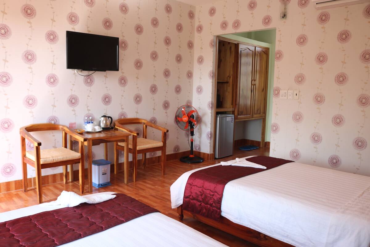 Ein Hotelzimmer mit zwei Betten, einem an der Wand befestigten Fernseher, einem kleinen Tisch mit Stühlen und einer Küchenzeile.