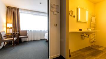 Habitació d'hotel amb un llit, una taula de treball amb una làmpada, una cadira, un bany amb un lavabo i un mirall, i una finestra amb cortines.