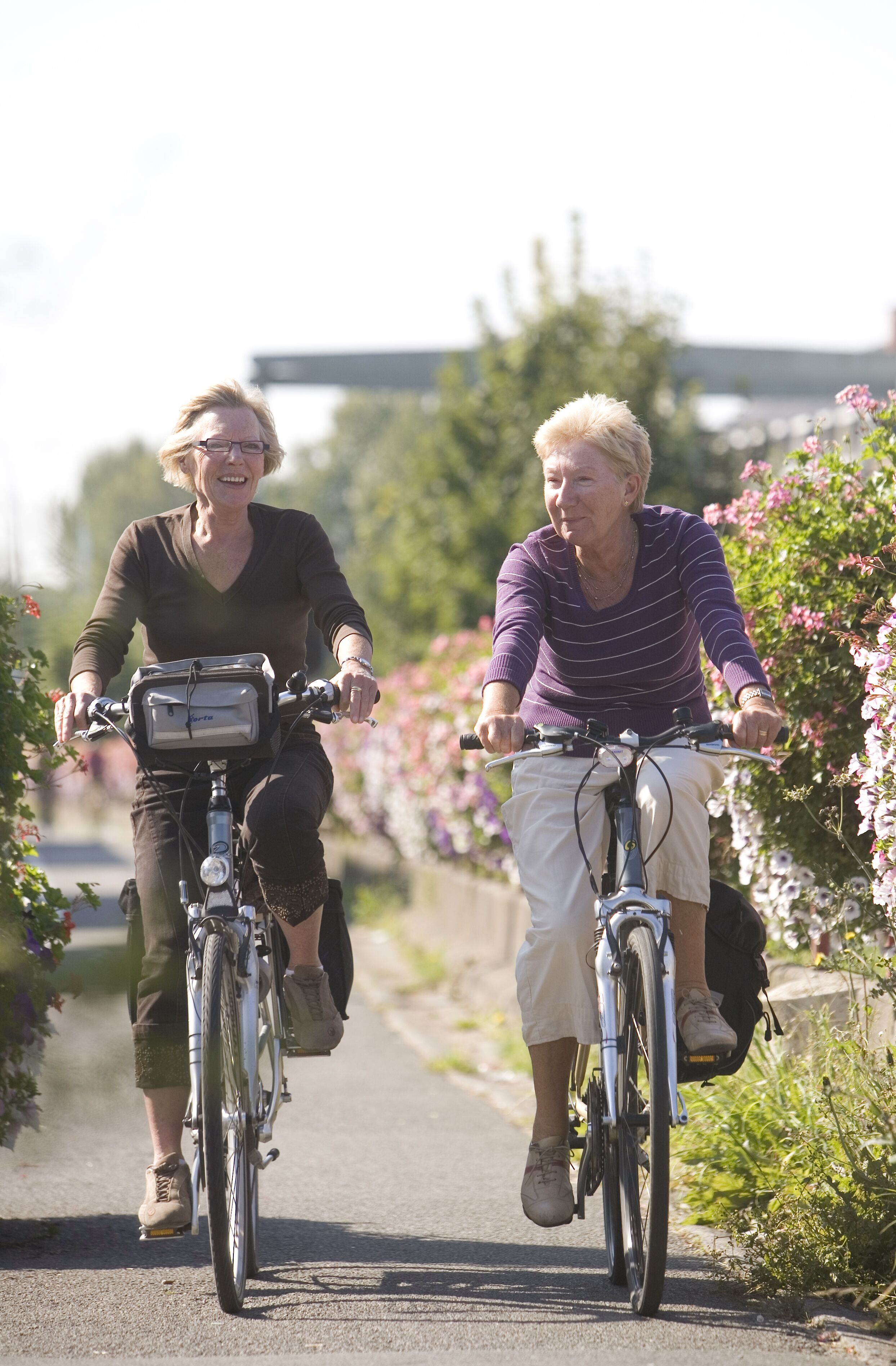 Two people riding bicycles on a paved path surrounded by greenery and flowers.