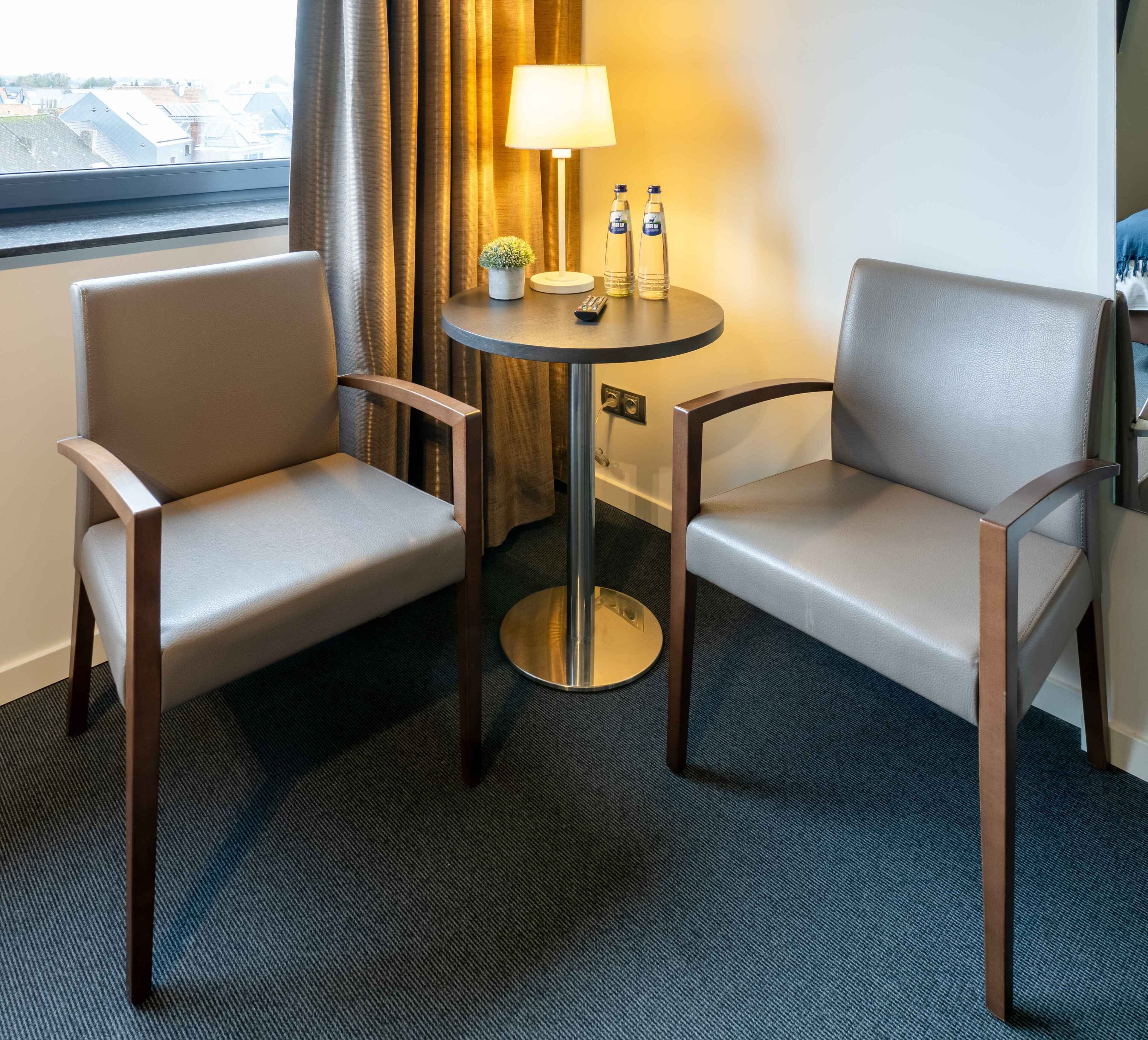 A hotel room with two armchairs, a small round table with a lamp, and a view of the city through a window.