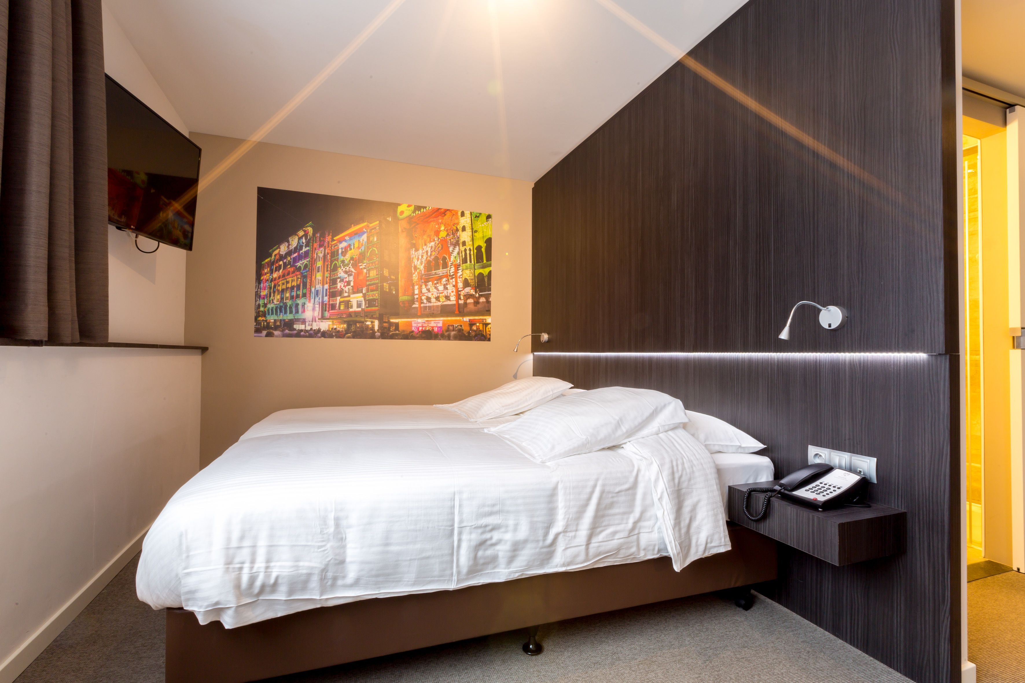 Une chambre d’hôtel moderne dotée d’un grand lit, d’un téléviseur à écran plat fixé au mur, d’un téléphone sur la table de chevet et d’un luminaire fixé au mur.