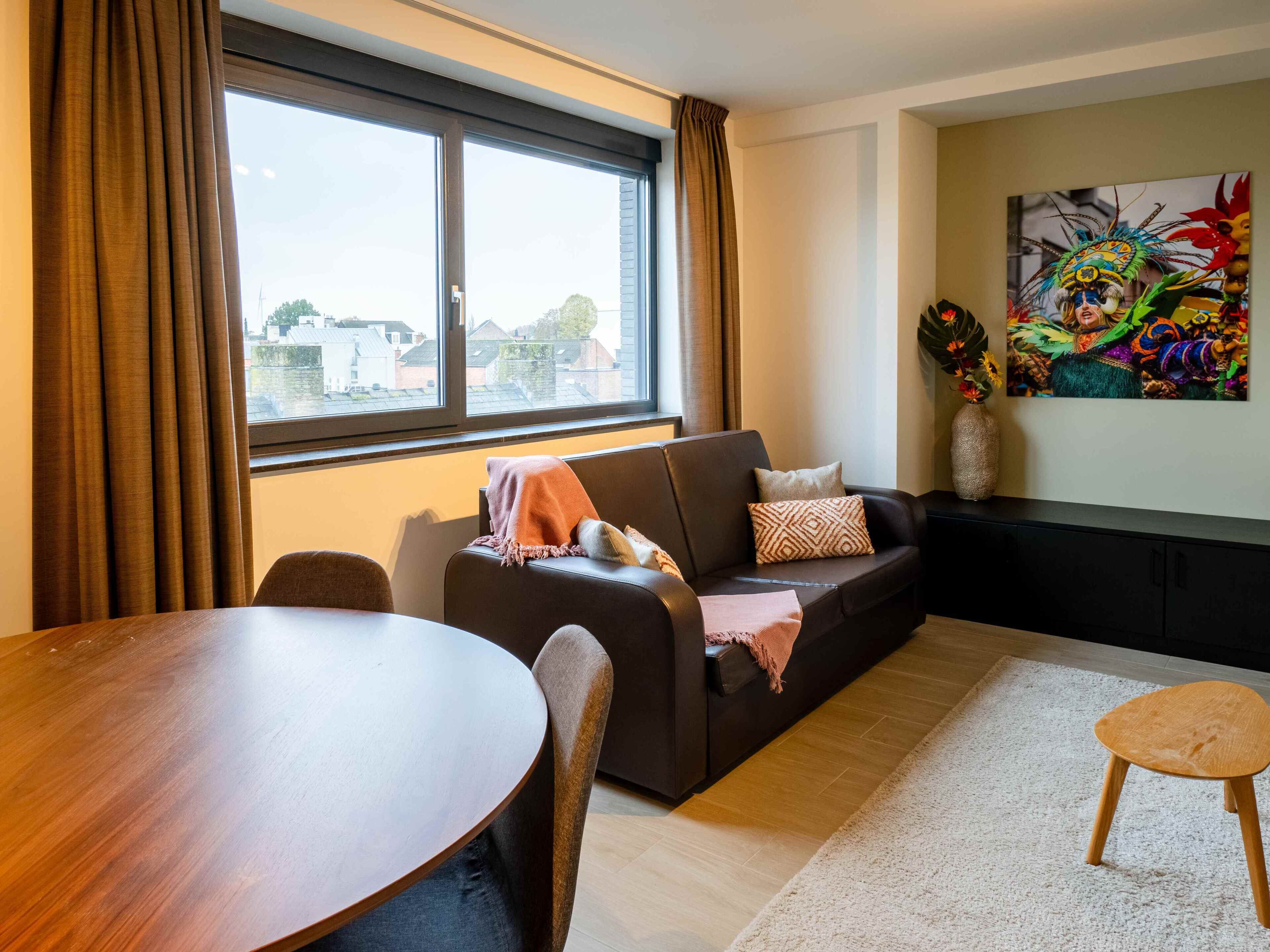 A modern living room with a dark brown sofa, a round wooden dining table, and a large window with curtains.