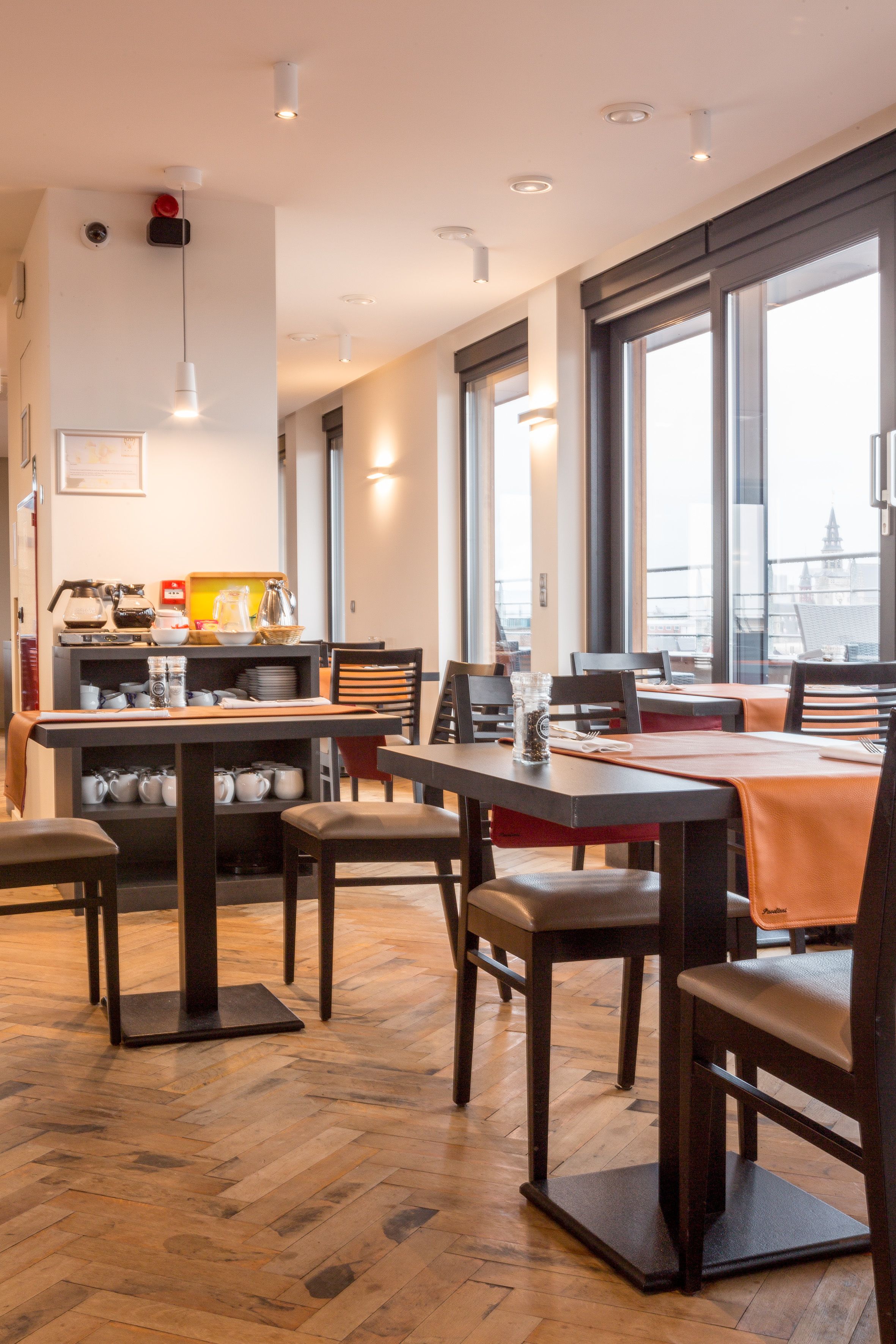A modern dining area with wooden flooring, tables, and chairs. There are large windows providing natural light and a view of the city.