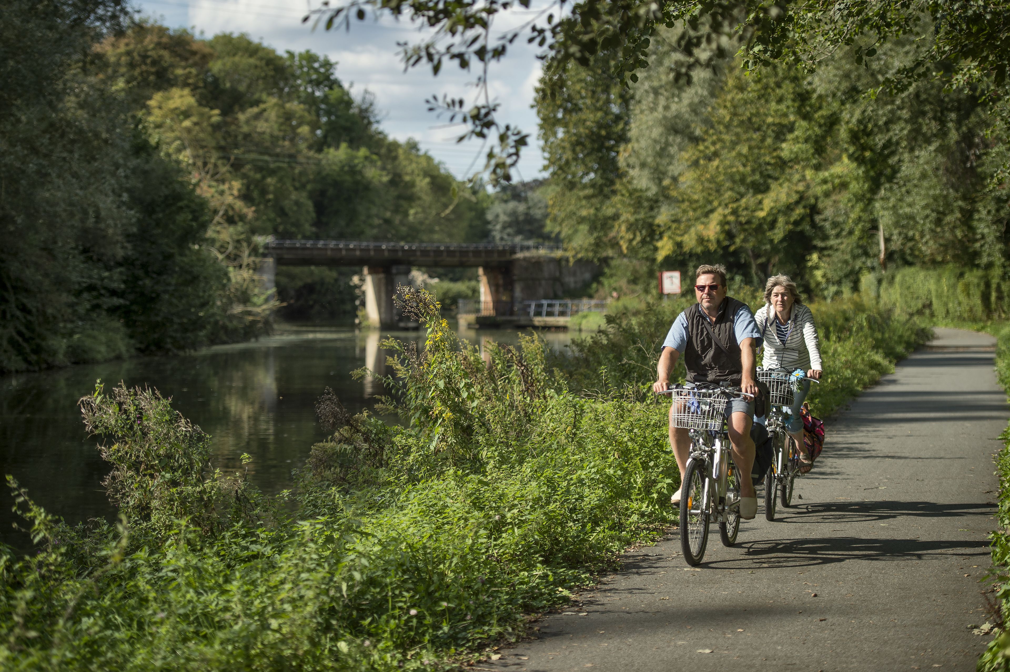 Un sentier longeant une rivière, avec des cyclistes et un pont en arrière-plan.