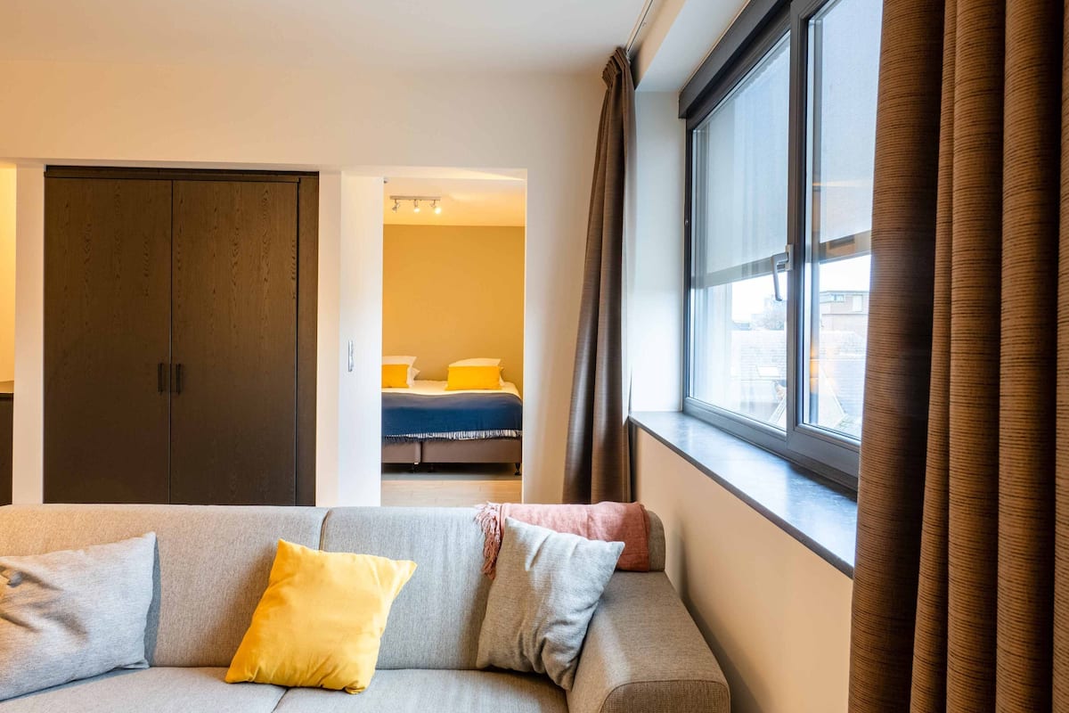 Una habitación de hotel moderna con un sofá gris, una cama con almohadas amarillas y un gran ventanal con cortinas.