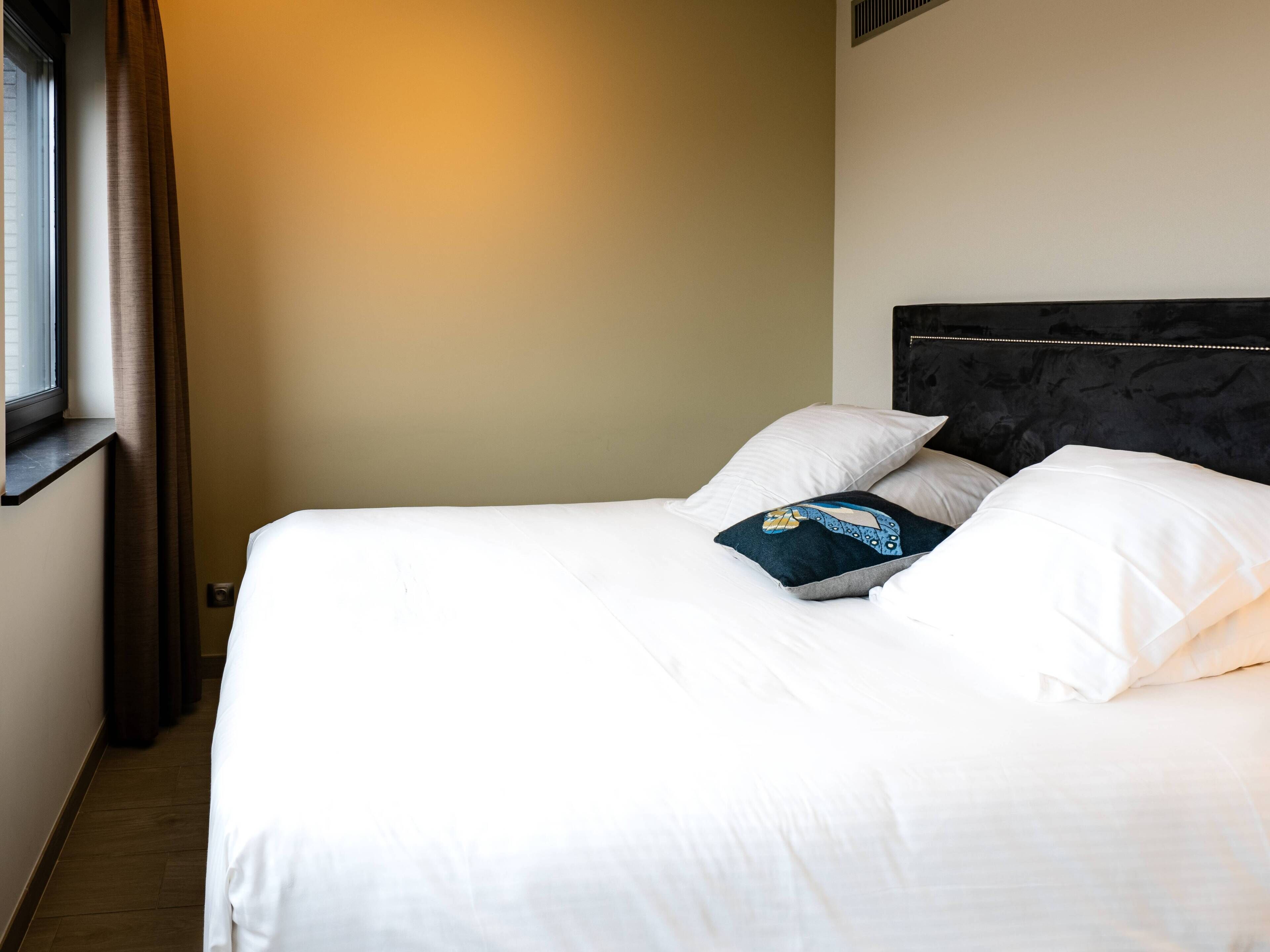 Una habitación de hotel con cama, ventana con cortinas y cabecero con lámpara.