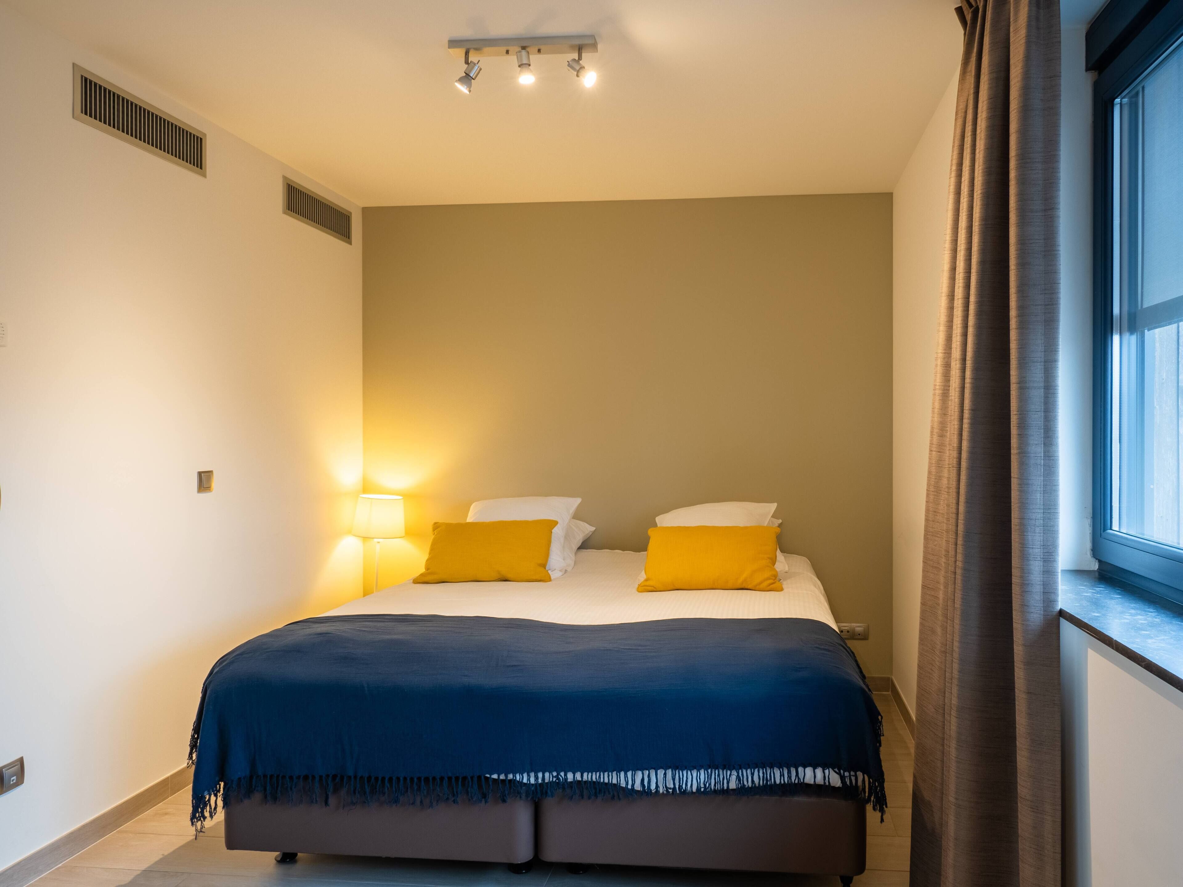 A bedroom with a bed, two bedside lamps, a window with curtains, and a ceiling light fixture.