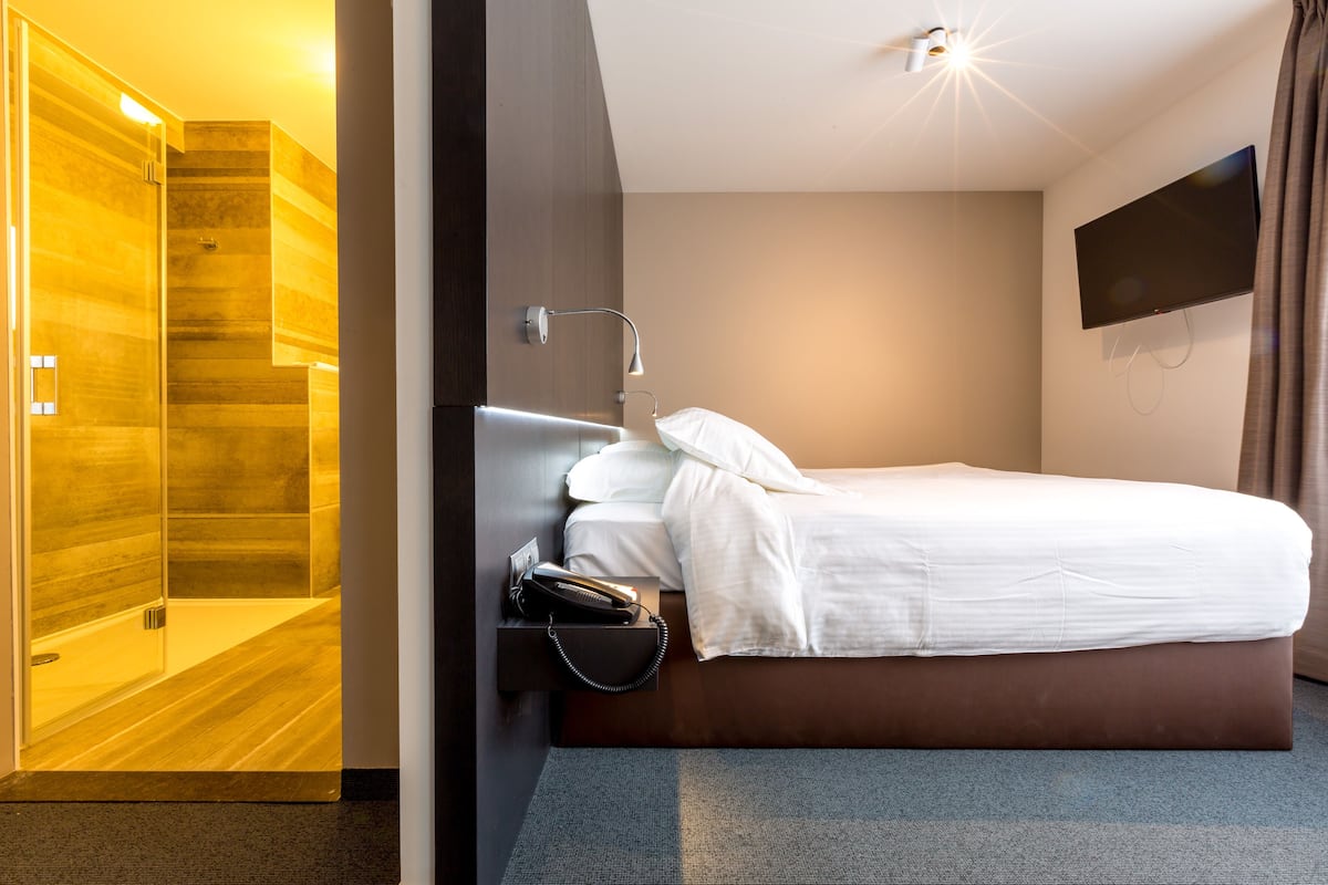 Una habitación de hotel moderna con suelo de madera, una cama con ropa blanca, una mesita de noche con lámpara, un televisor montado en la pared y una zona de ducha.