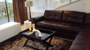 A modern living room with a black leather sofa, a white ottoman, and a black coffee table with magazines and candles. - Villa La Vida- Self-Catering House (Umhlanga)
