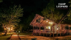 Een rustieke, rieten dakbedekte constructie met een houten terras, omringd door bomen en een grindpad bij nacht.