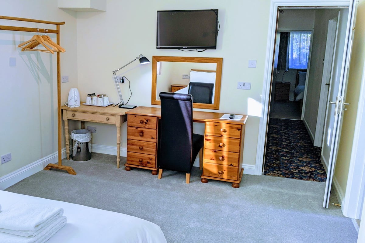 A hotel room with a bed, a desk with a chair, a dresser, a mirror, a television, and a hanging coat rack.
