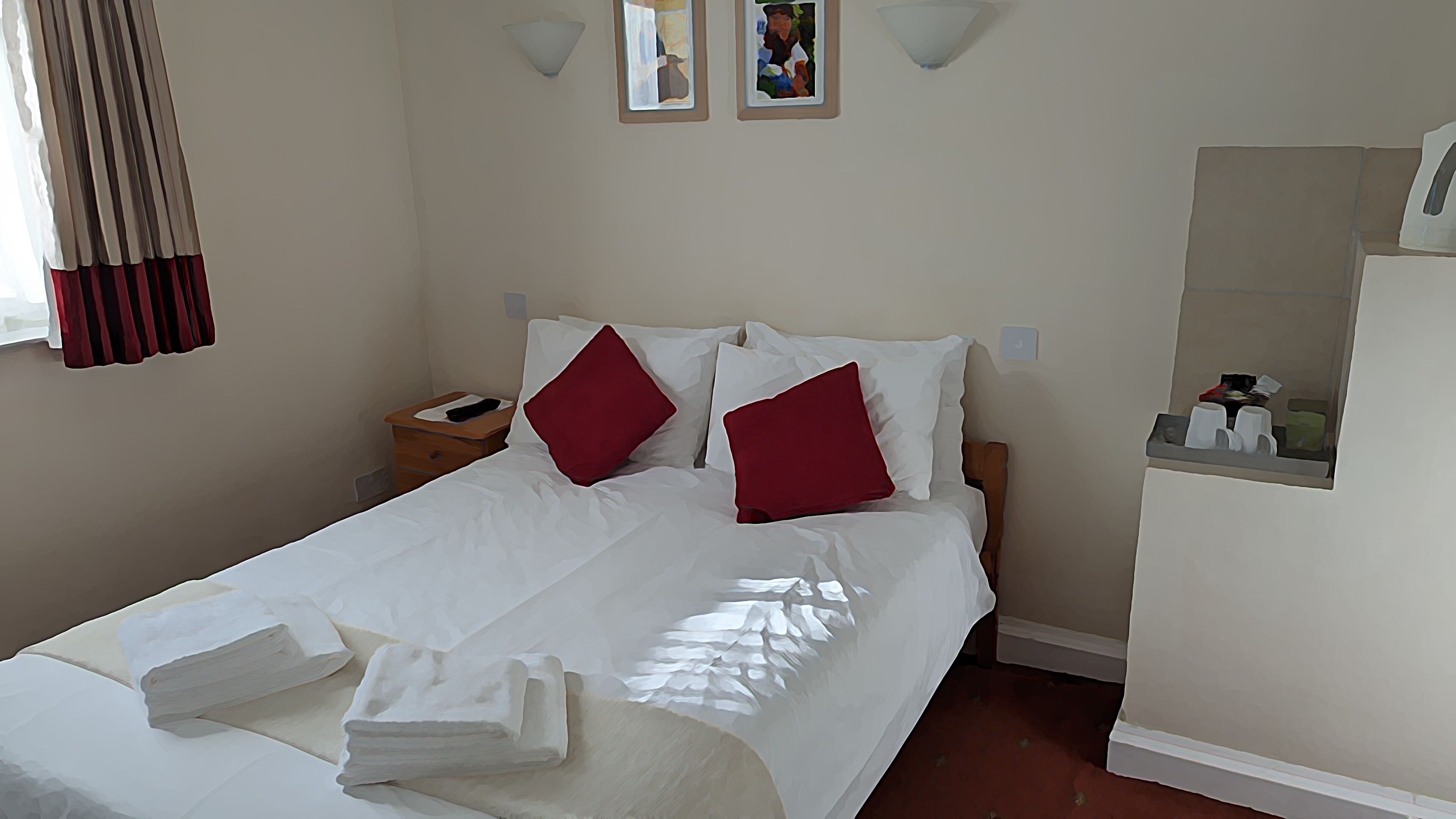 Un lit bien fait, avec du linge de lit blanc et deux oreillers rouges, une table de chevet en bois sur laquelle se trouve un objet noir, un luminaire fixé au mur et une petite étagère avec des articles de toilette.
