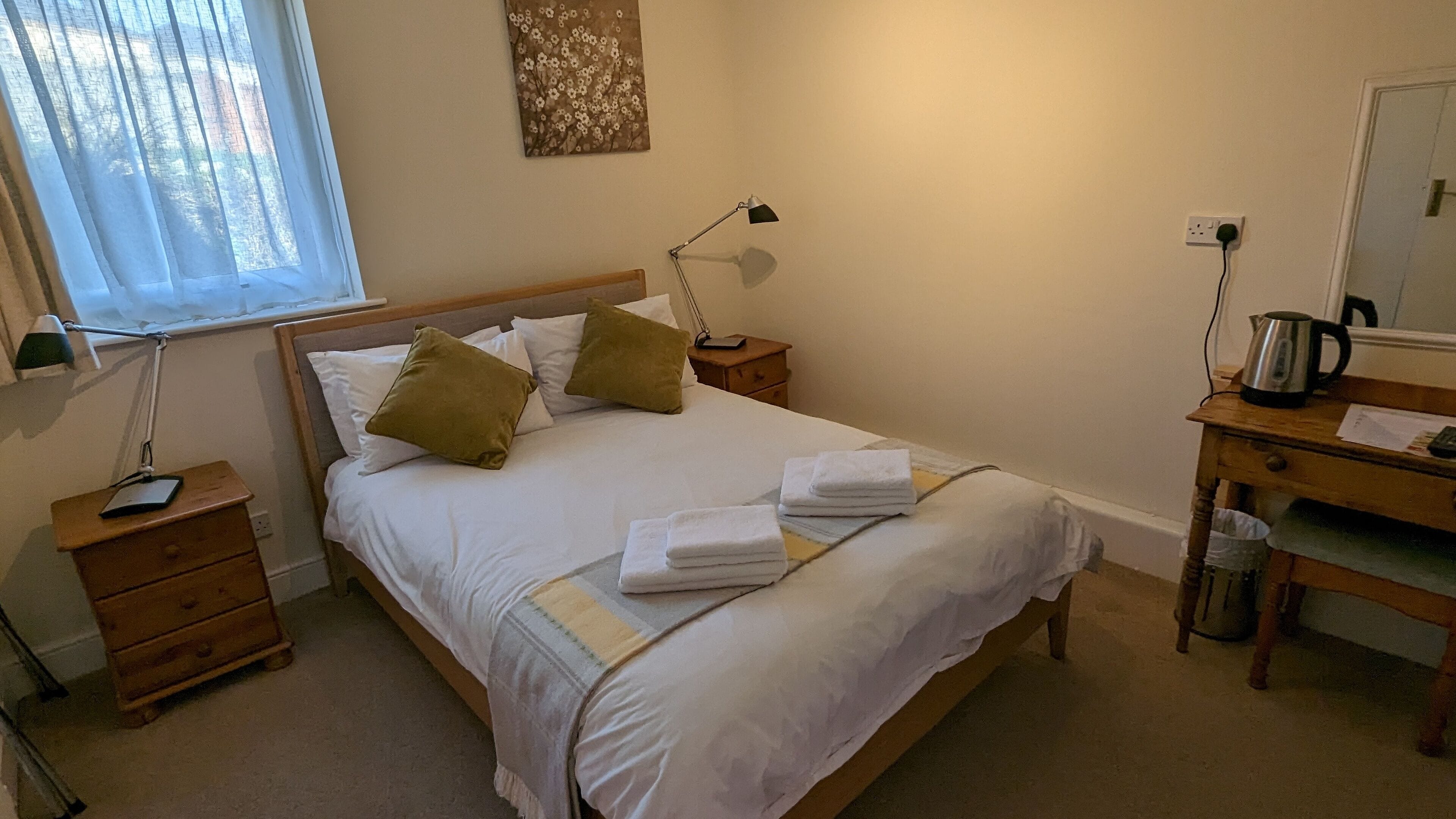Een keurig opgemaakt bed met kussens, een bedlampje, een houten nachtkastje, een klein ladeblok en een bureau met waterkoker en papieren.