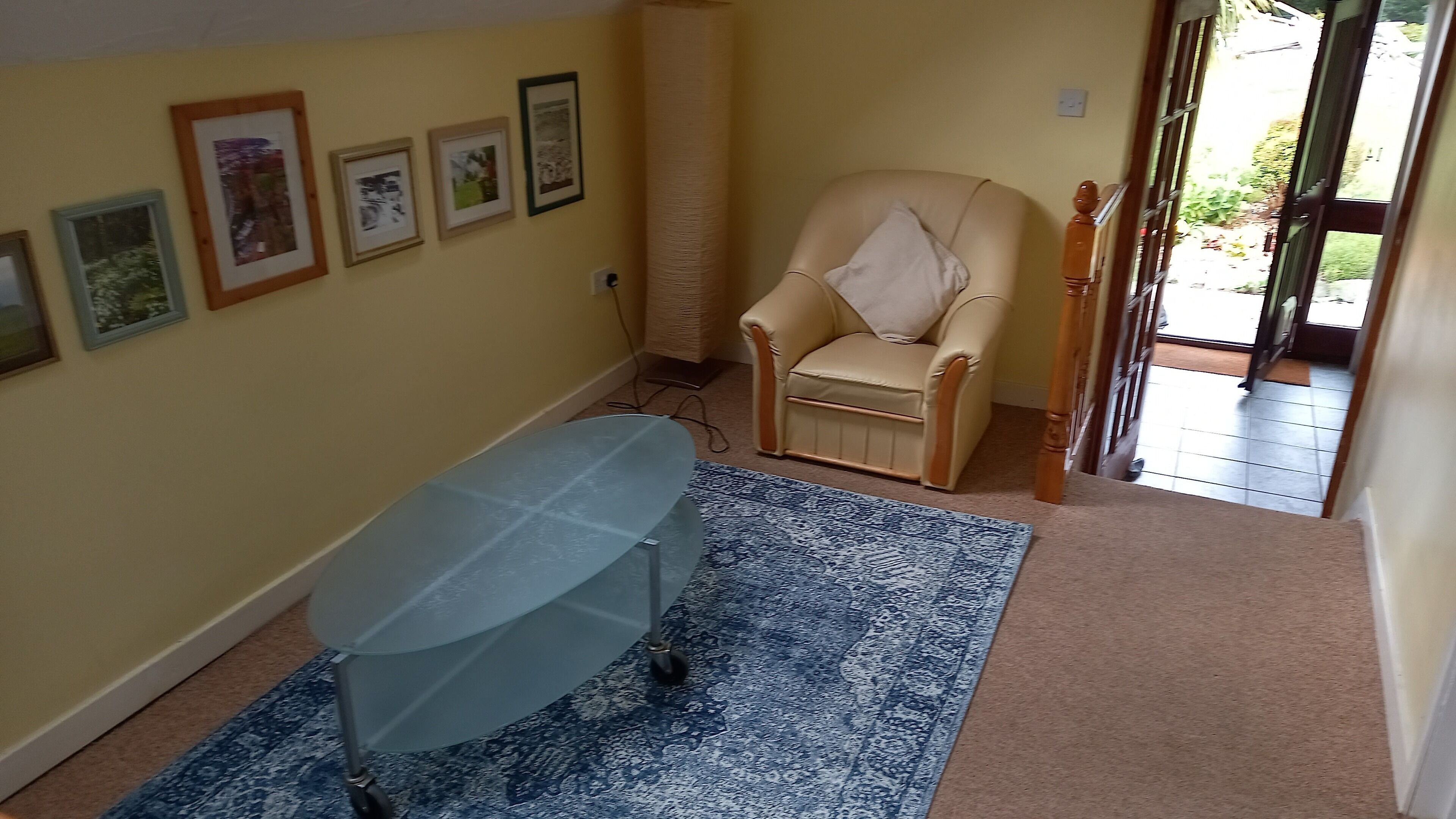 Una habitación con un tapete azul estampado, una mesa con cubierta de vidrio, un sillón color beige y varias fotografías enmarcadas en la pared.