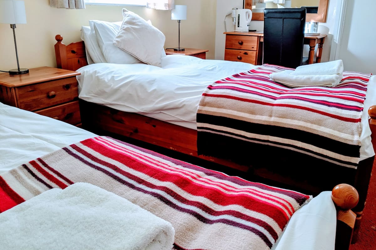 A hotel room with two beds, a wooden dresser, a nightstand, a lamp, a mirror, and a window with curtains.