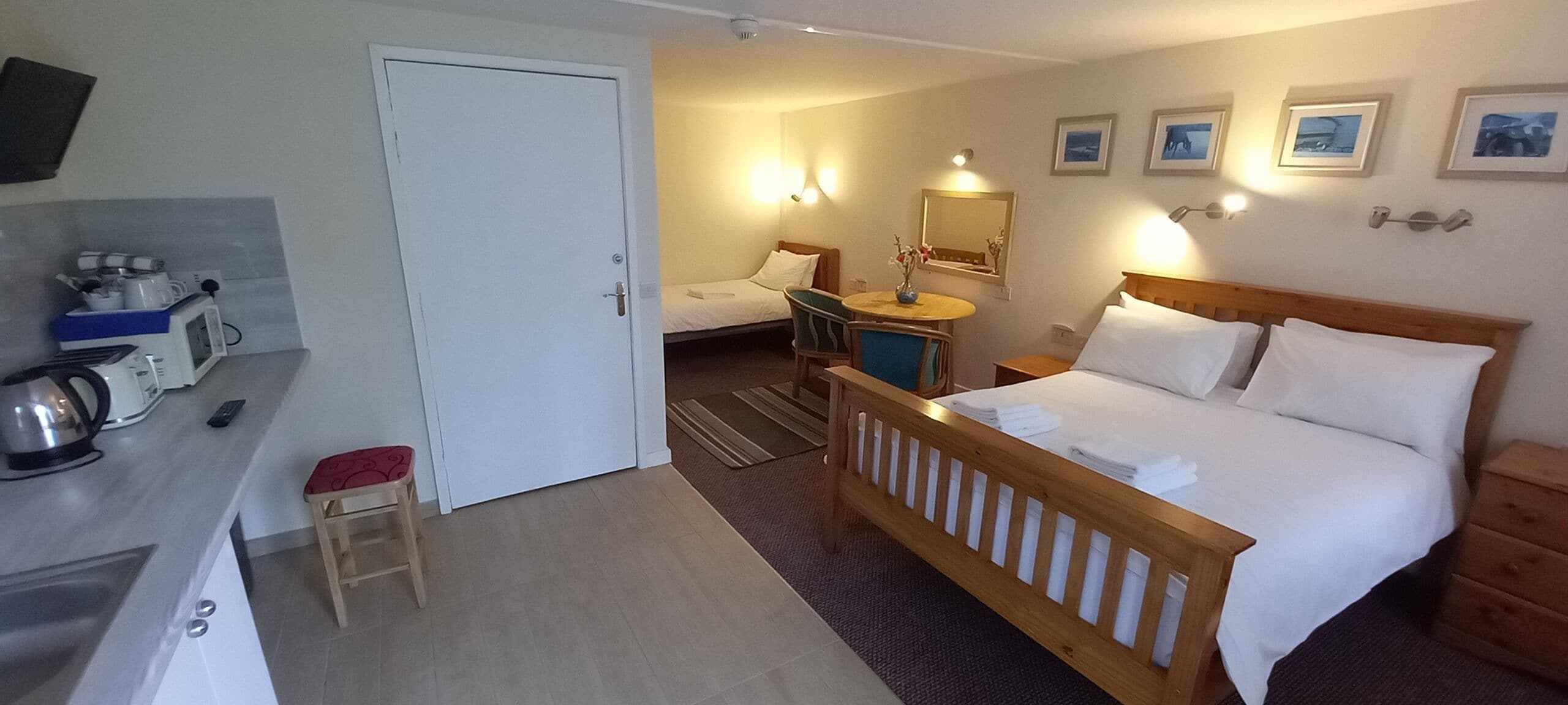 A hotel room with a bed, a desk with a kettle, a chair, a small table with a vase, and a door.
