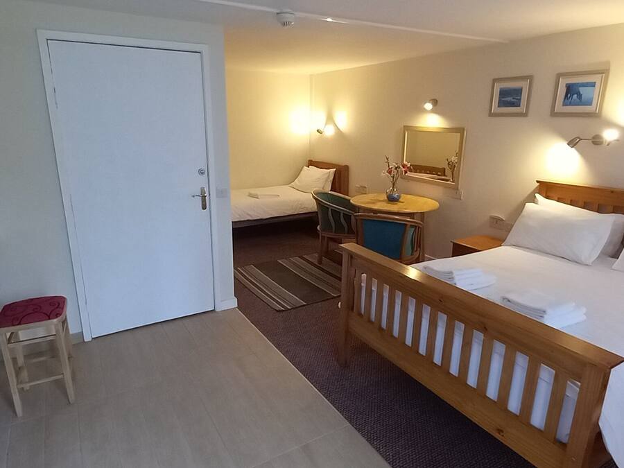 A hotel room with a bed, a desk with a kettle, a chair, a small table with a vase, and a door.