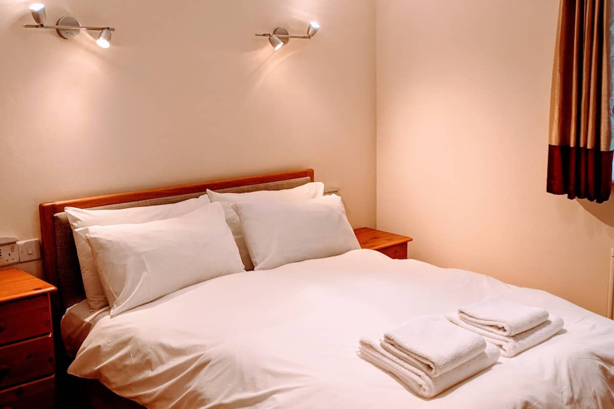 A neatly made bed with white linens and towels, a wooden headboard, bedside tables, and wall-mounted lighting.