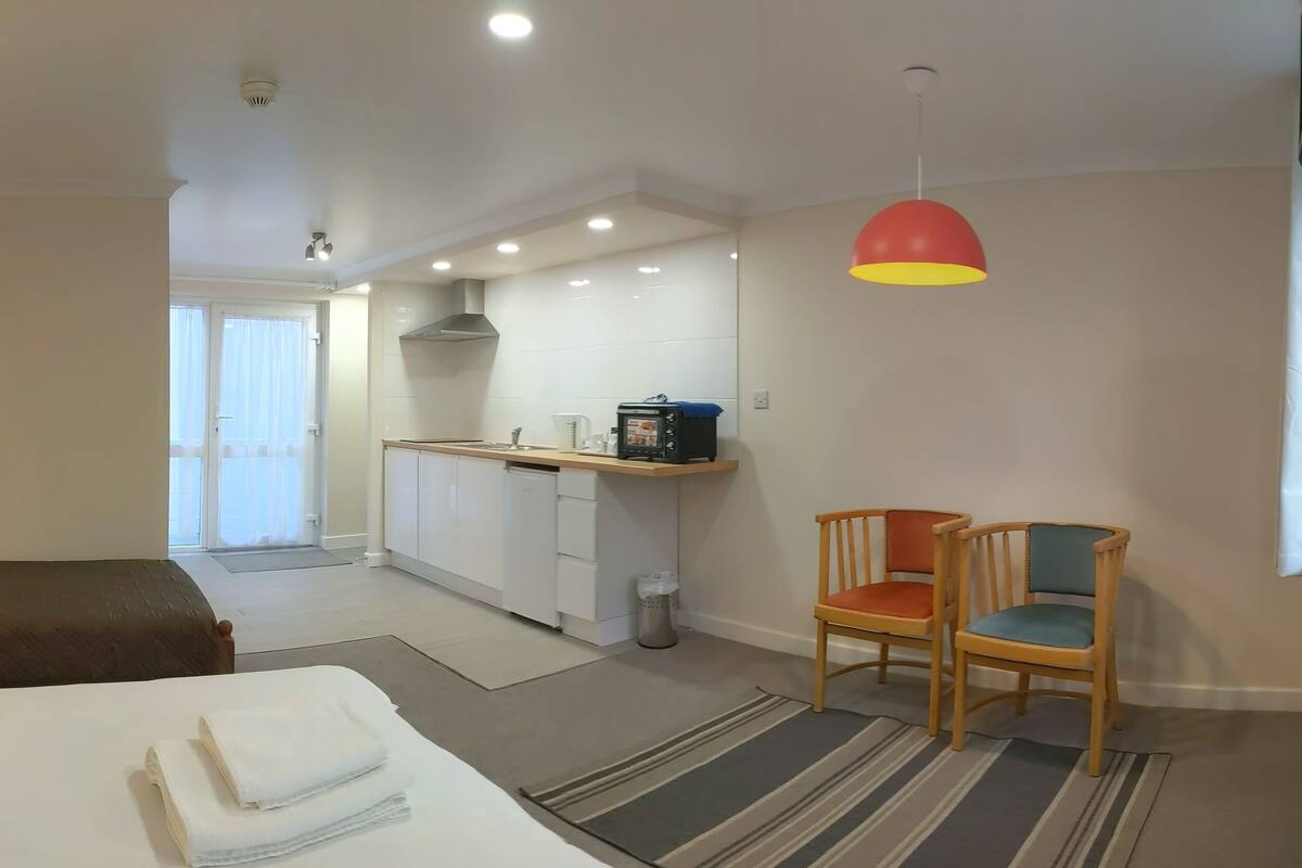 A modern hotel room with a kitchenette, a dining area with two chairs, a bed with neatly folded towels, and a striped rug.