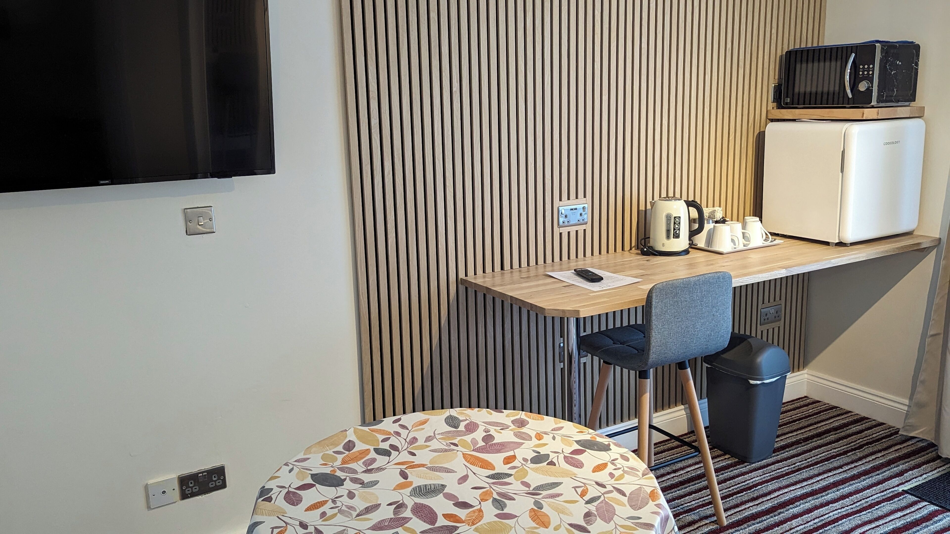 A compact room with a wooden desk, a chair, a small table, a microwave, a refrigerator, and a television.