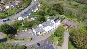 An aerial view of a residential area with a prominent white house featuring a large garage and solar panels, surrounded by lush greenery and a curved road.