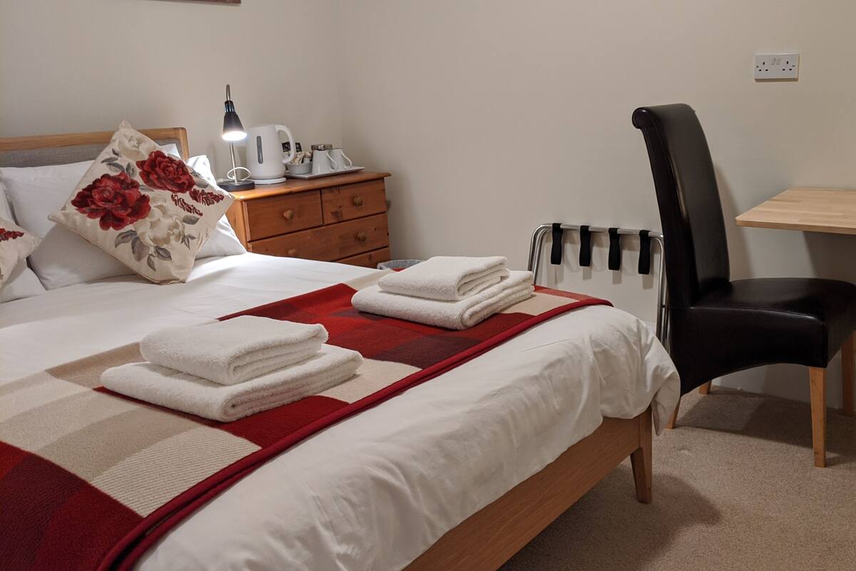 A neatly made bed with white linens and a red and white patterned blanket. There is a wooden nightstand with a white kettle and cups, a black chair, and a wooden desk with a lamp.