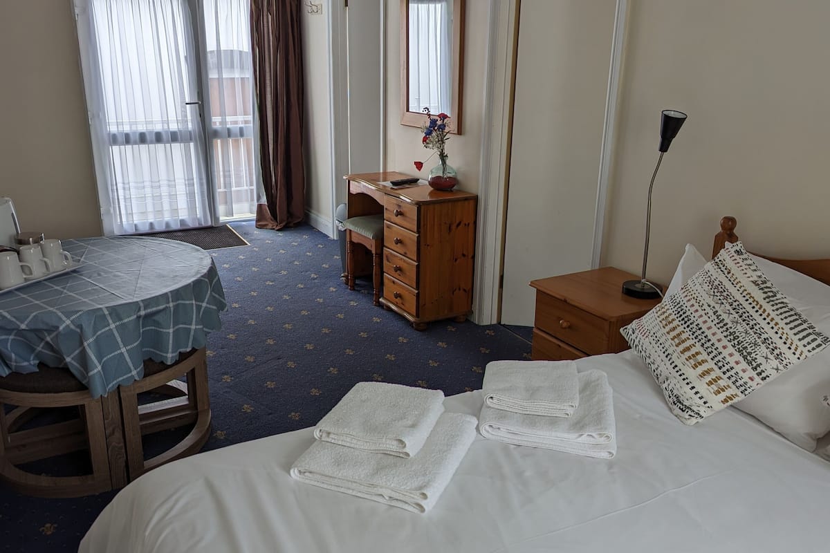 A hotel room with a bed, a desk with a chair, a dresser, a bedside table, and a window with curtains.