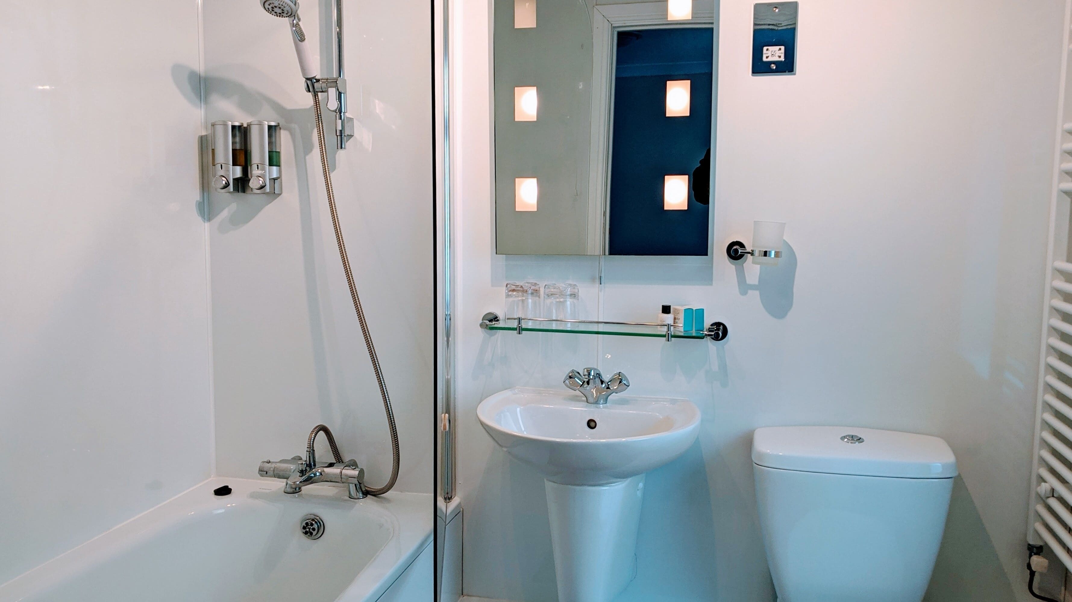 Ein Badezimmer mit einer Badewanne, einem Waschbecken und einer Toilette. Über der Badewanne befindet sich ein Duschkopf, über dem Waschbecken ein Spiegel.
