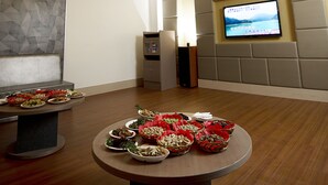 Uma sala de conferências moderna com uma mesa redonda exibindo diversos pratos, uma tela grande na parede e um design de teto singular.