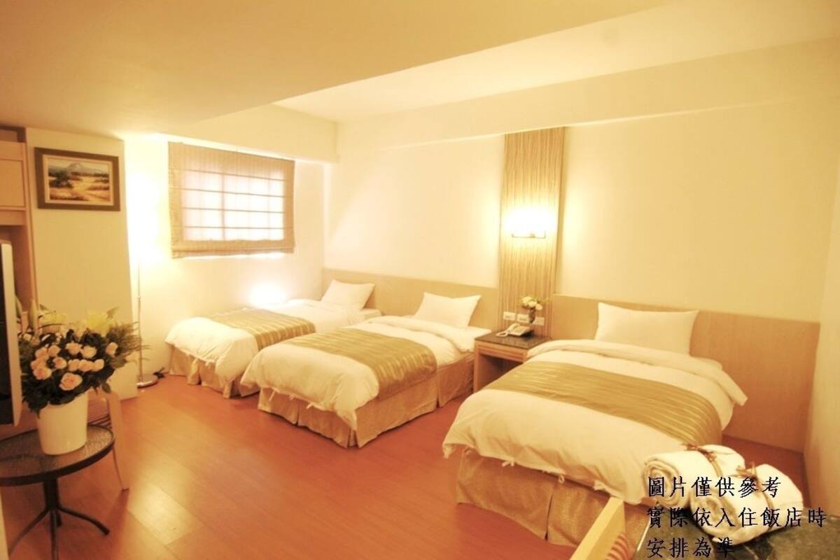 A hotel room with two beds, a small table with a vase of flowers, a bedside table with a lamp, and a window with blinds.