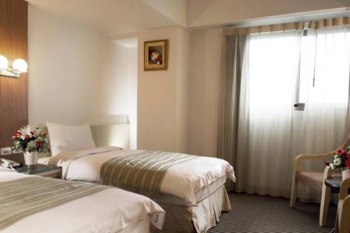 A hotel room with two beds, a nightstand with a phone, a chair, a table with a vase of flowers, a painting on the wall, and a window with curtains.