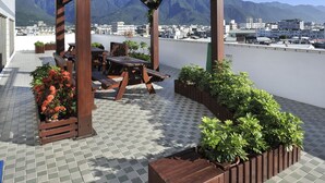 Uma pérgola de madeira com assentos e vasos em um pátio ladrilhado, com vista para um porto e montanhas.
