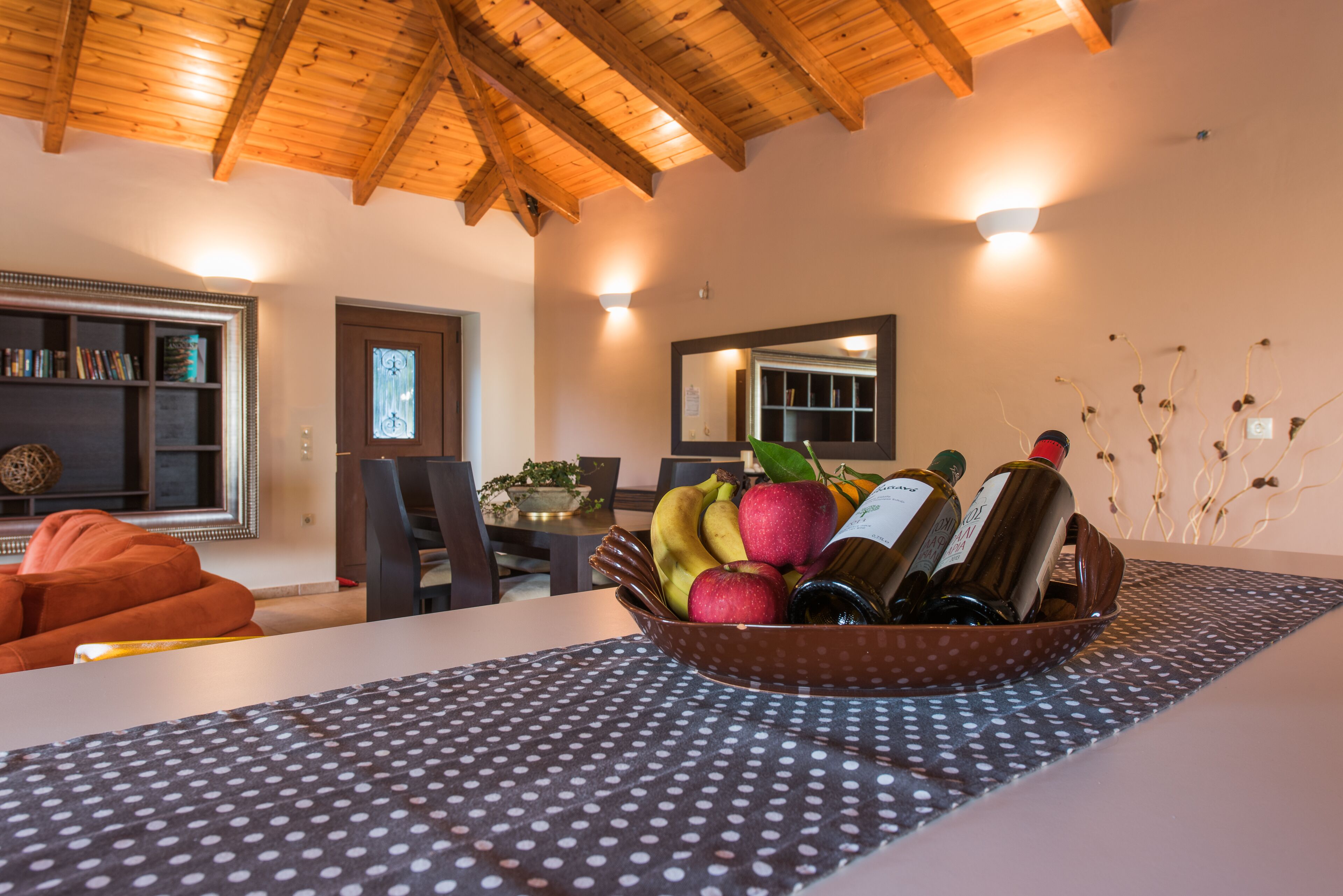 Un coin repas avec un plafond en bois, une table recouverte d’une nappe à pois et un saladier rempli de fruits, ainsi que des bouteilles de vin.