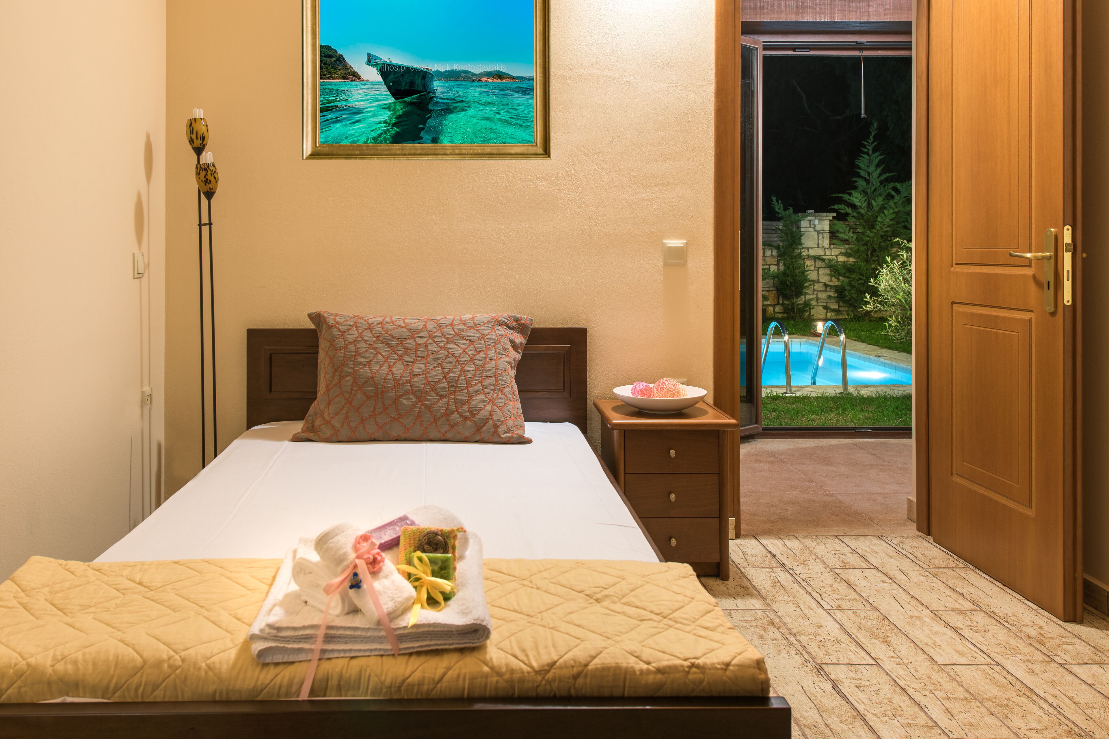A hotel room with a bed, a nightstand, a framed picture of a bridge, and a view of an outdoor swimming pool.