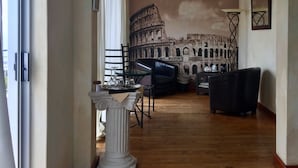 A living room with a large mural of the Colosseum, a glass-top table with a tray, a black leather armchair, and a white pedestal. - Villa Surprise (Cape Town)