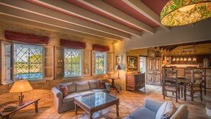 Amplia sala de estar con sofá, sillones y una mesa de centro. Se observan grandes ventanales con rejas de hierro decorativas y cortinas rojas. En el fondo, se distingue una zona de cocina con armarios de madera y una mesa de comedor.