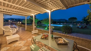 Un patio techado con mobiliario exterior, una mesa de vidrio con fruta y aperitivos, y vistas a una piscina y jardines al anochecer.
