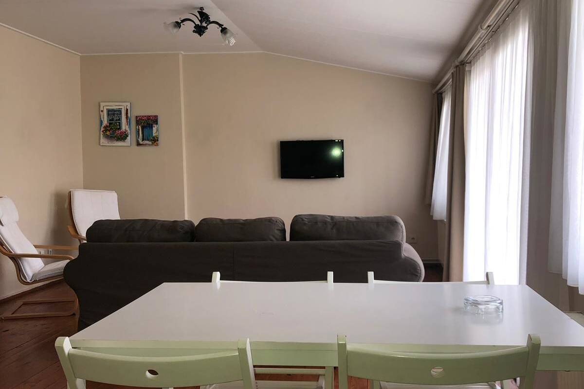 A living room with a white table and four green chairs, a gray sofa, a mounted TV, and a ceiling-mounted light fixture.