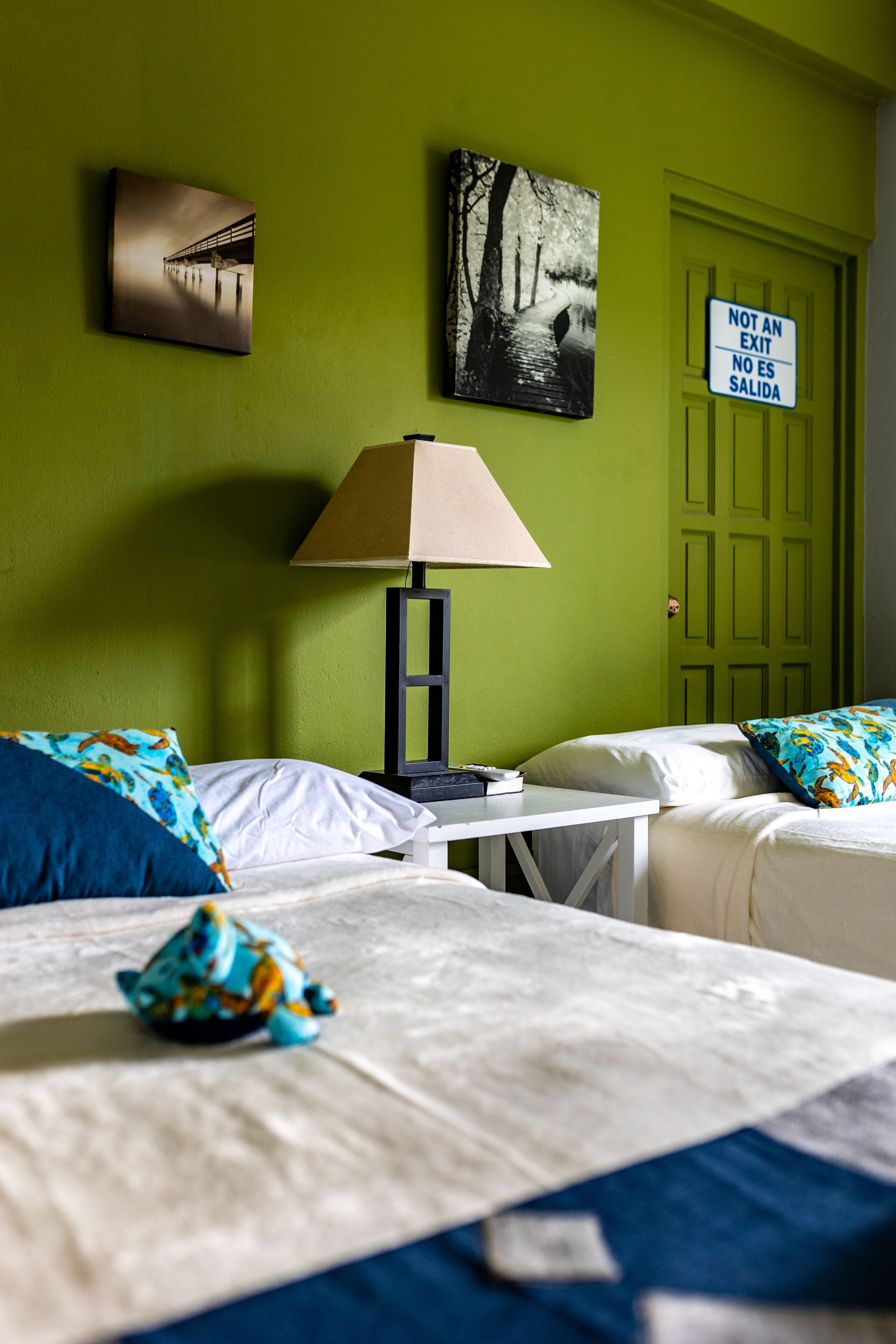 A hotel room with two beds, a nightstand, a lamp, and a green door with a sign.