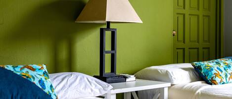 A hotel room with two beds, a nightstand, a lamp, and a green door with a sign.