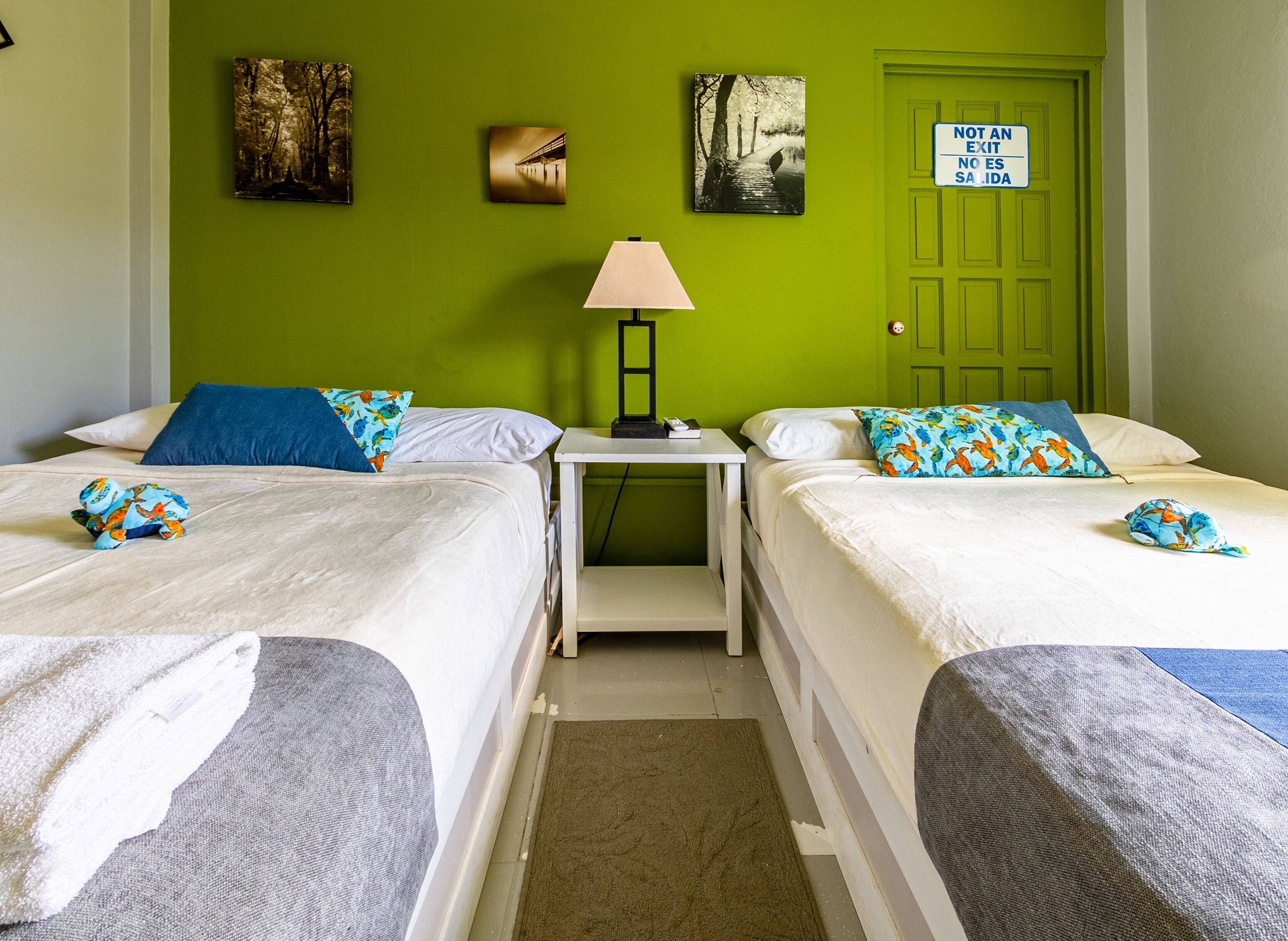 A room with two beds, a nightstand with a lamp, and a green door with a sign.