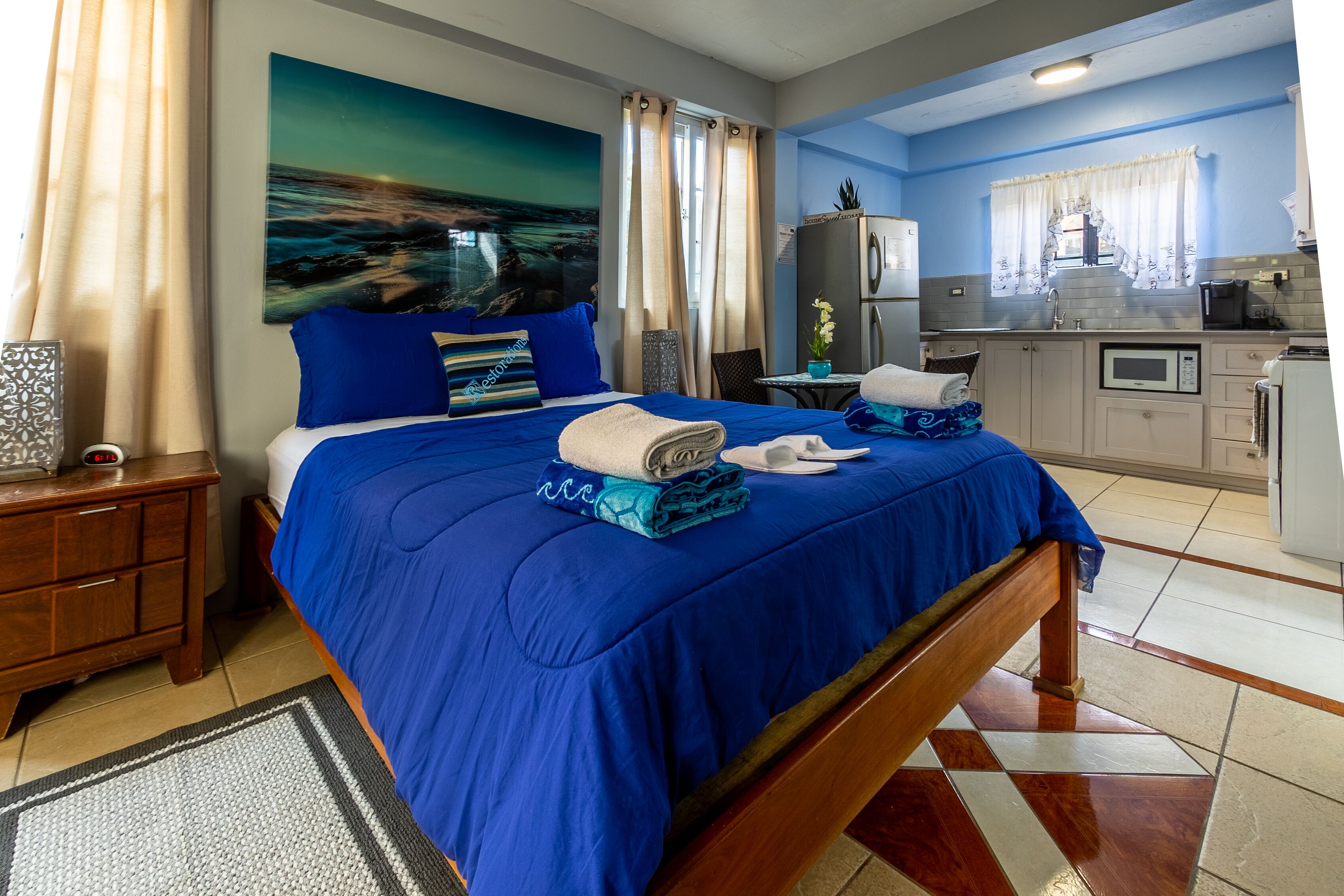 A hotel room with a bed, a nightstand, a desk, a window with curtains, a kitchen area with a refrigerator, microwave, and sink.