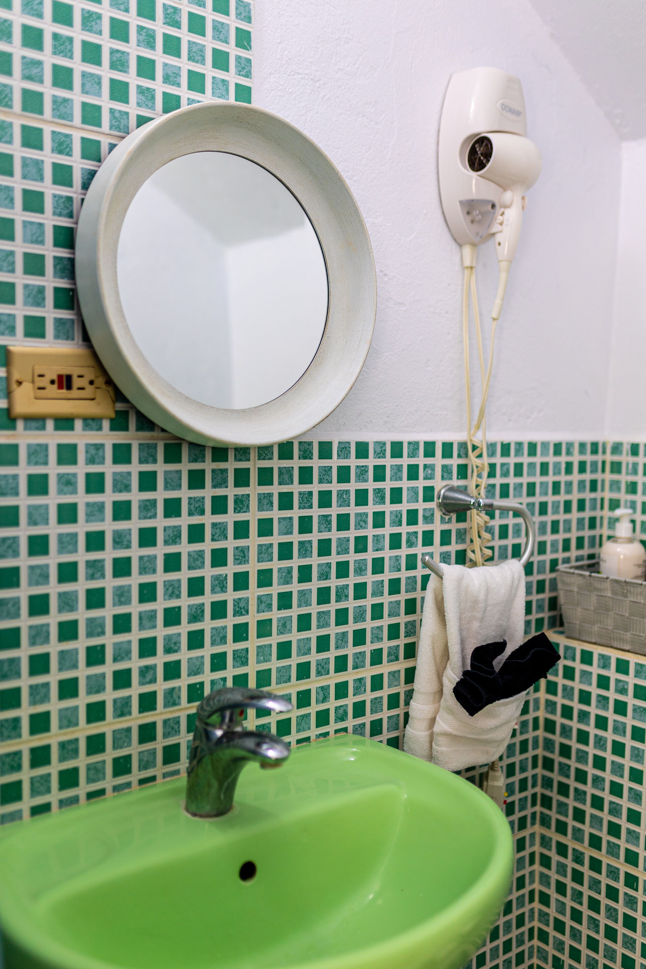 A bathroom with a green sink, a round mirror, a hairdryer, and a towel rack.