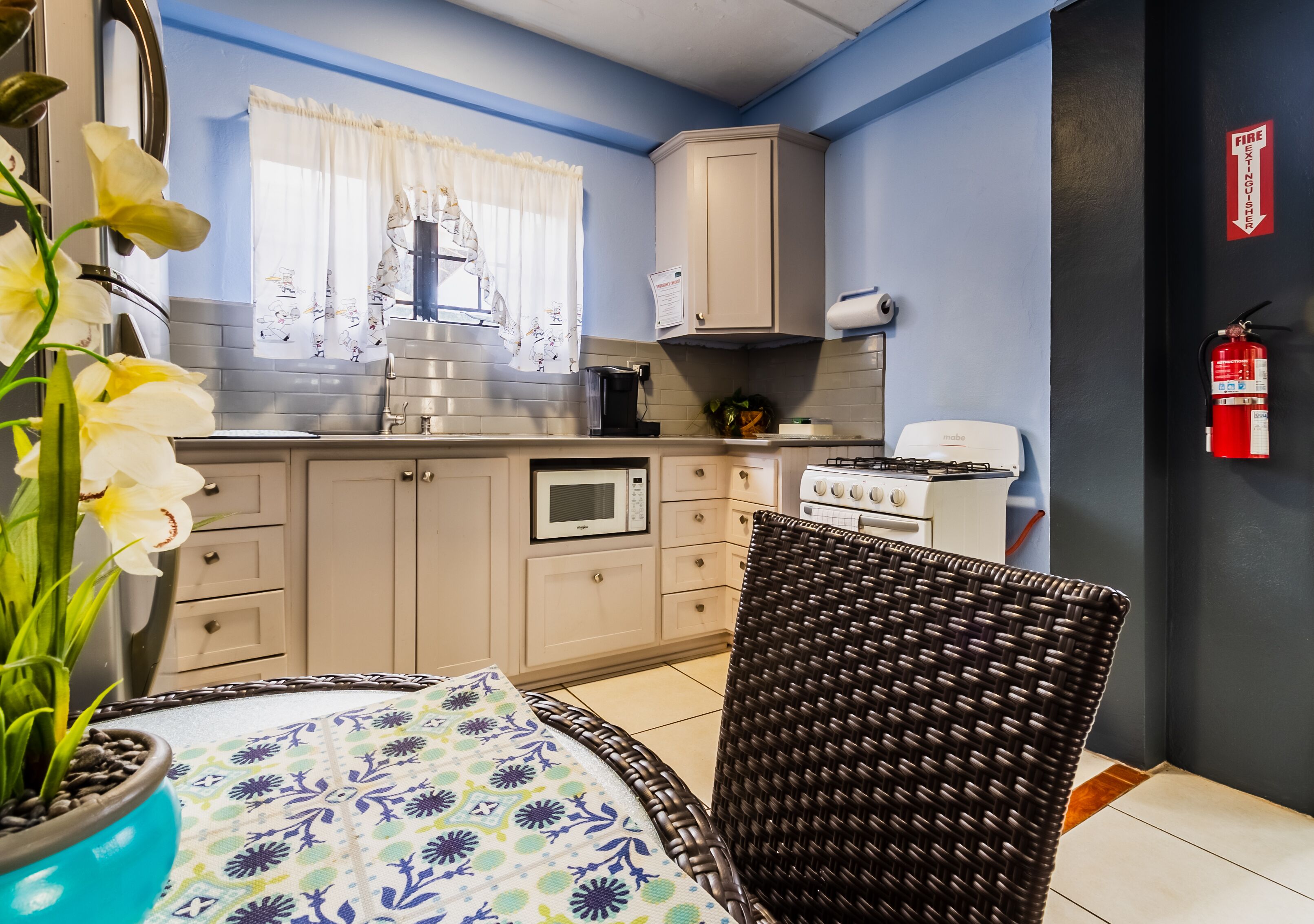 A compact kitchen with a microwave, stove, and cabinets, a wicker chair, and a potted plant.
